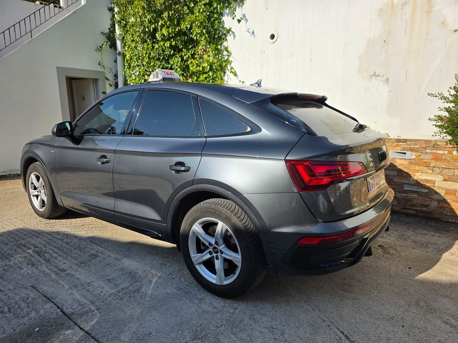 Une Audi Q5 Sportback gris foncé est garée sur une allée pavée à côté d'un bâtiment blanc.