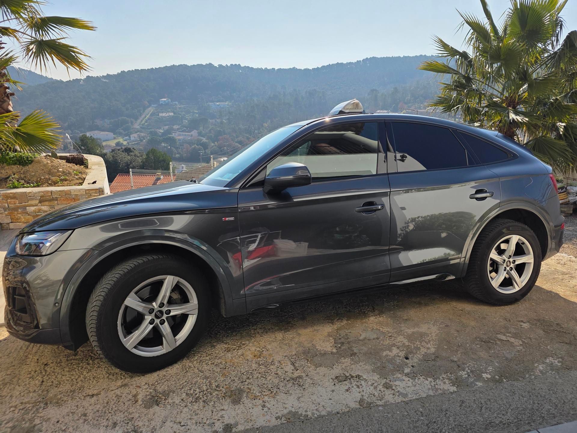 Une Audi Q5 Sportback gris foncé garée sur un terrain en terre battue, avec des palmiers et une colline en arrière-plan.