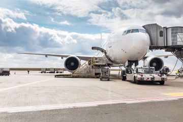 Vue grand angle d'un avion de ligne blanc stationné à une porte d'embarquement d'aéroport, pris en charge par des équipements et des véhicules au sol.