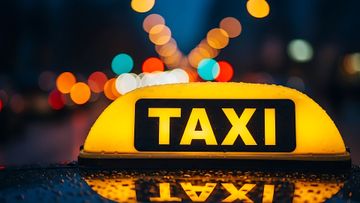 Le gyrophare jaune vif d'un taxi brille dans l'obscurité, sur un fond flou de lumières urbaines colorées et de reflets de surface.