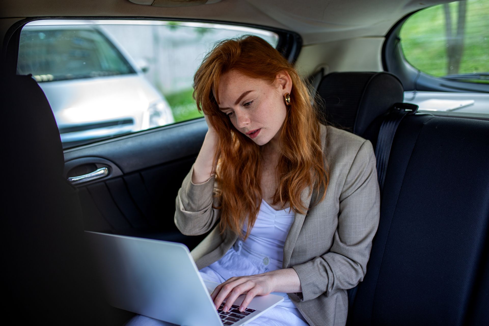 Une personne aux cheveux roux, vêtue d'un blazer, est assise à l'arrière d'une voiture, le regard baissé, en train de taper sur un ordinateur portable.