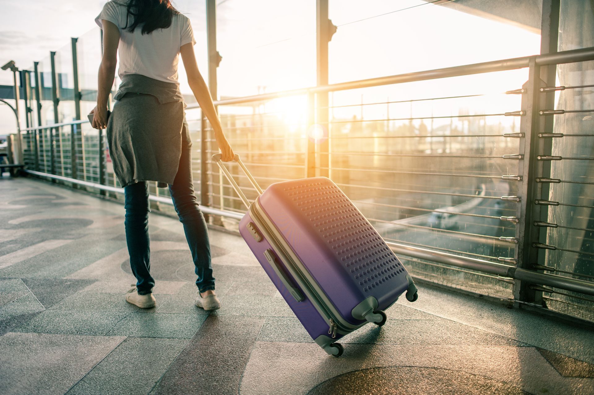 Au coucher du soleil, une personne marche sur une allée en tirant derrière elle une valise à roulettes violette.