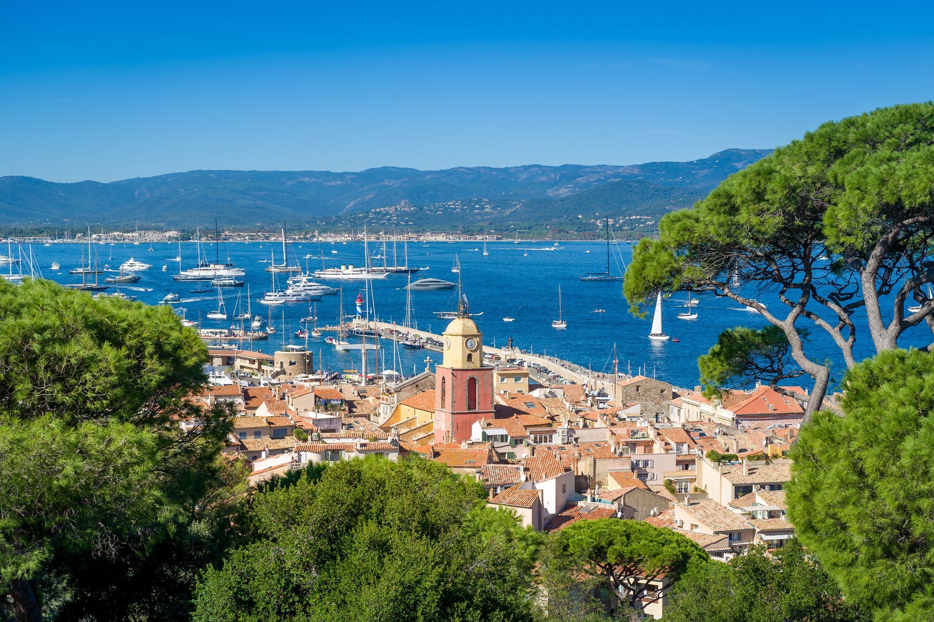 Une ville portuaire méditerranéenne ensoleillée, avec des bâtiments aux toits rouges, une tour d'horloge imposante et de nombreux bateaux sur une eau bleue.