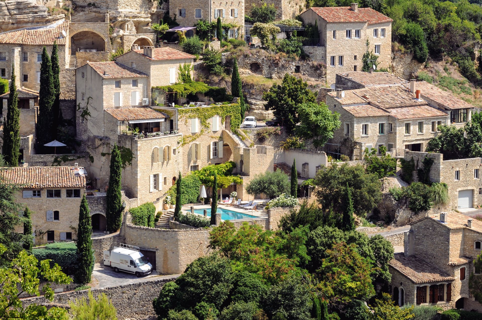 Un village de pierre ensoleillé en France, avec ses maisons rustiques, sa végétation luxuriante et sa petite piscine nichée entre les bâtiments.