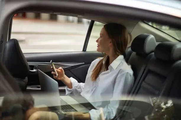 Une personne vêtue d'une chemise blanche est assise à l'arrière d'une voiture, regardant par la fenêtre tout en tenant un smartphone et en utilisant un ordinateur portable.