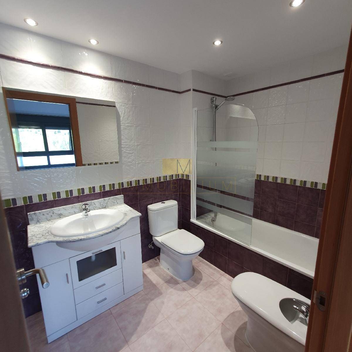 Baño con lavabo, inodoro y bañera blancos. Azulejos granates y blancos con mampara de cristal en la ducha.