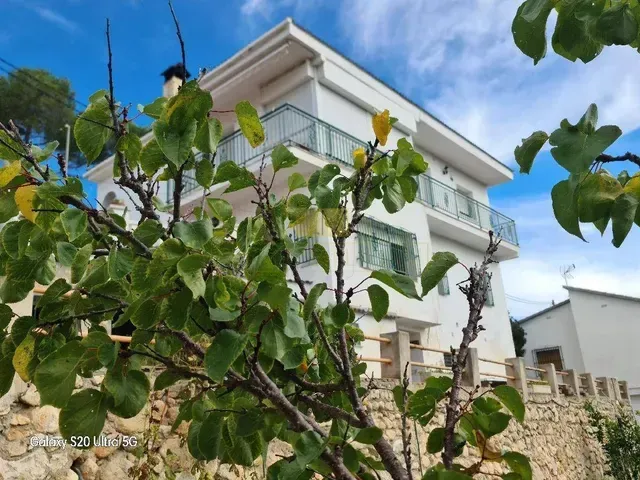 Casa blanca de dos pisos con balcones, tras frondosos árboles. Cielo azul con nubes.