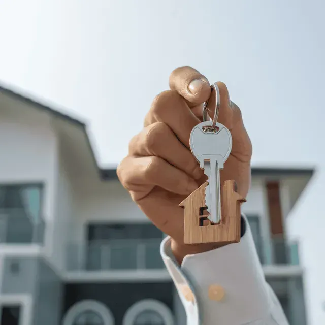 Mano sosteniendo llaves con llavero en forma de casa frente a una casa.