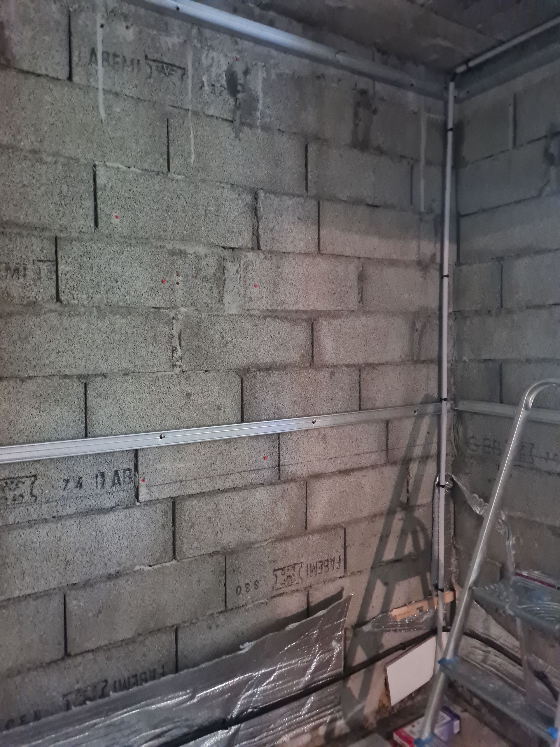 Murs en parpaings gris avec conduits électriques, une petite échelle et un sol partiellement recouvert.