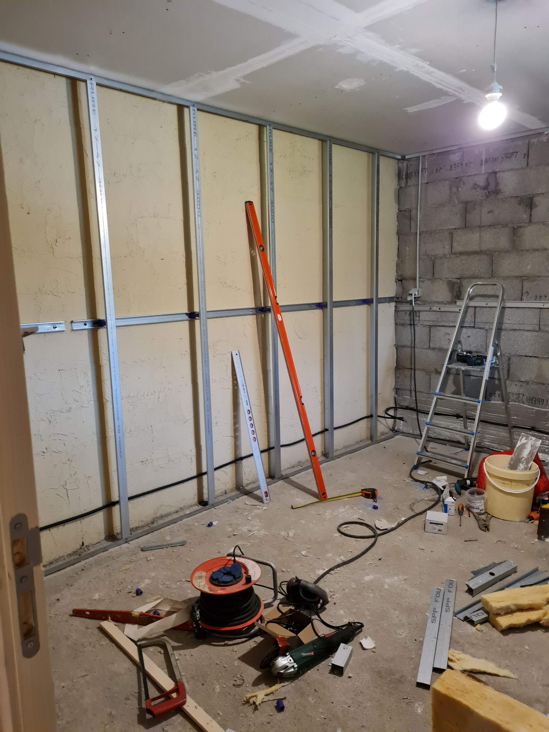 Intérieur d'une pièce en construction avec des montants métalliques, de l'isolant, des outils et une échelle adossée à un mur en parpaings.