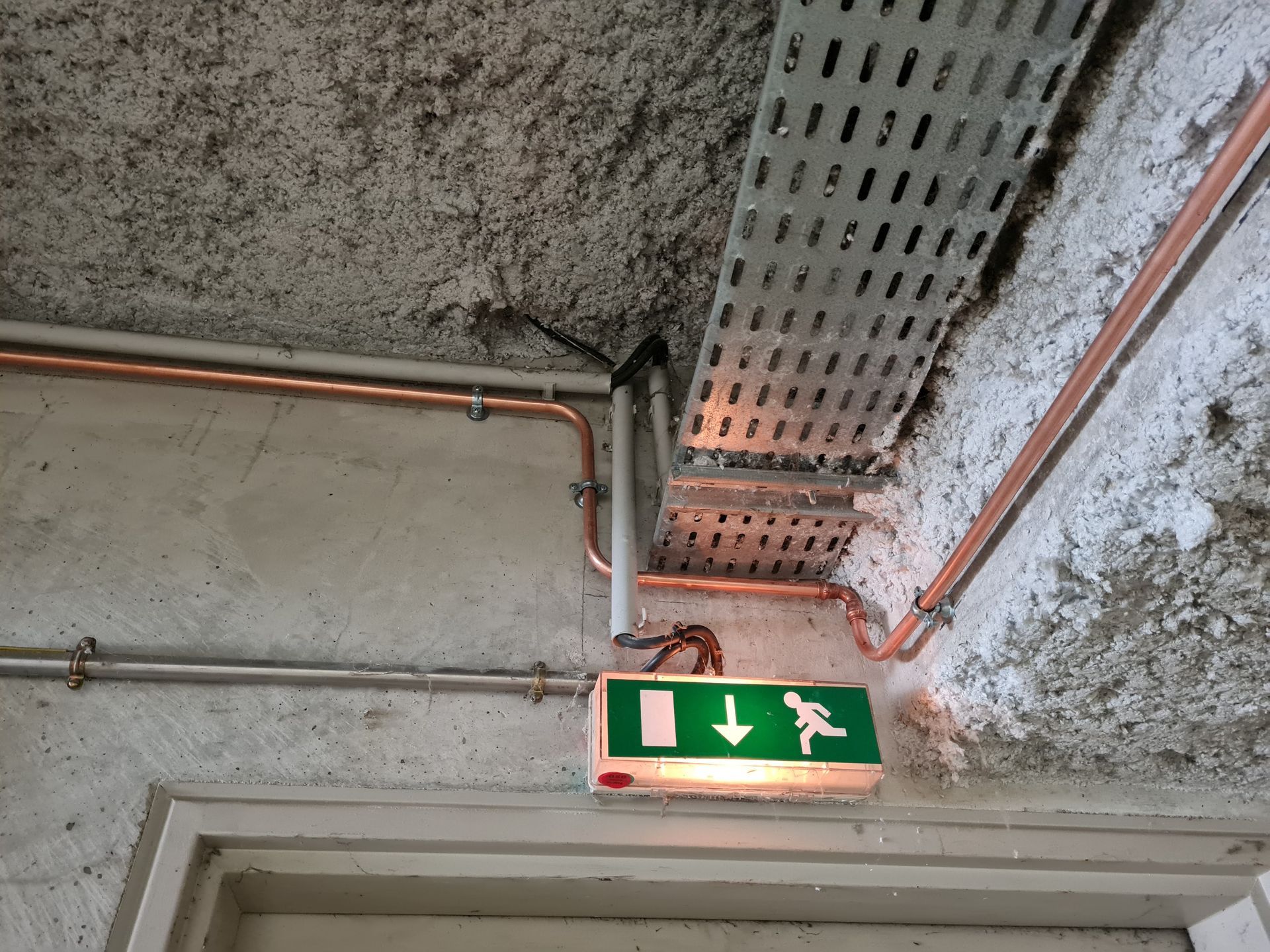 Panneau de sortie de secours avec flèche verte lumineuse, sous des tuyaux et un chemin de câbles métallique sur un plafond en béton.