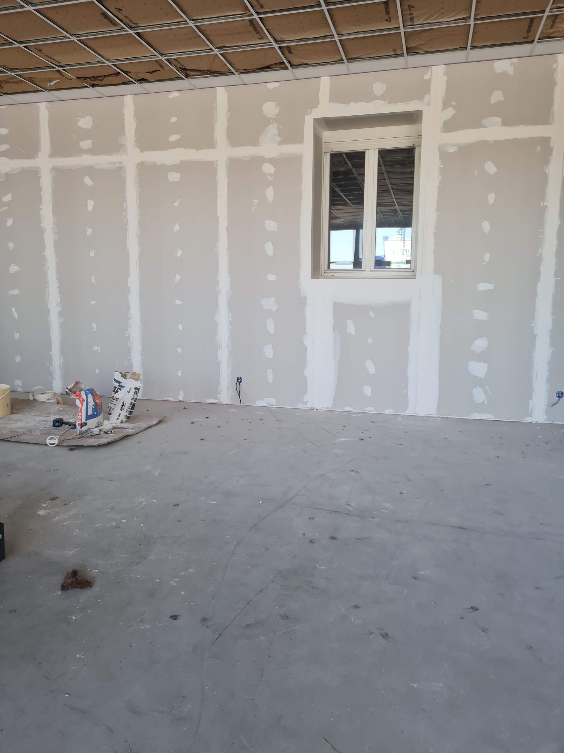 Pièce intérieure en cours d'aménagement, avec cloisons sèches et fenêtre. Sol en ciment et plafond apparent.