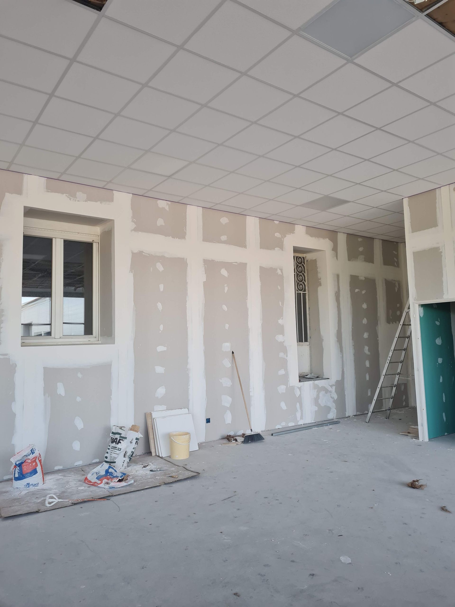 Chantier de construction intérieur avec murs et plafond en plaques de plâtre ; présence d'une fenêtre, d'une échelle et d'outils.