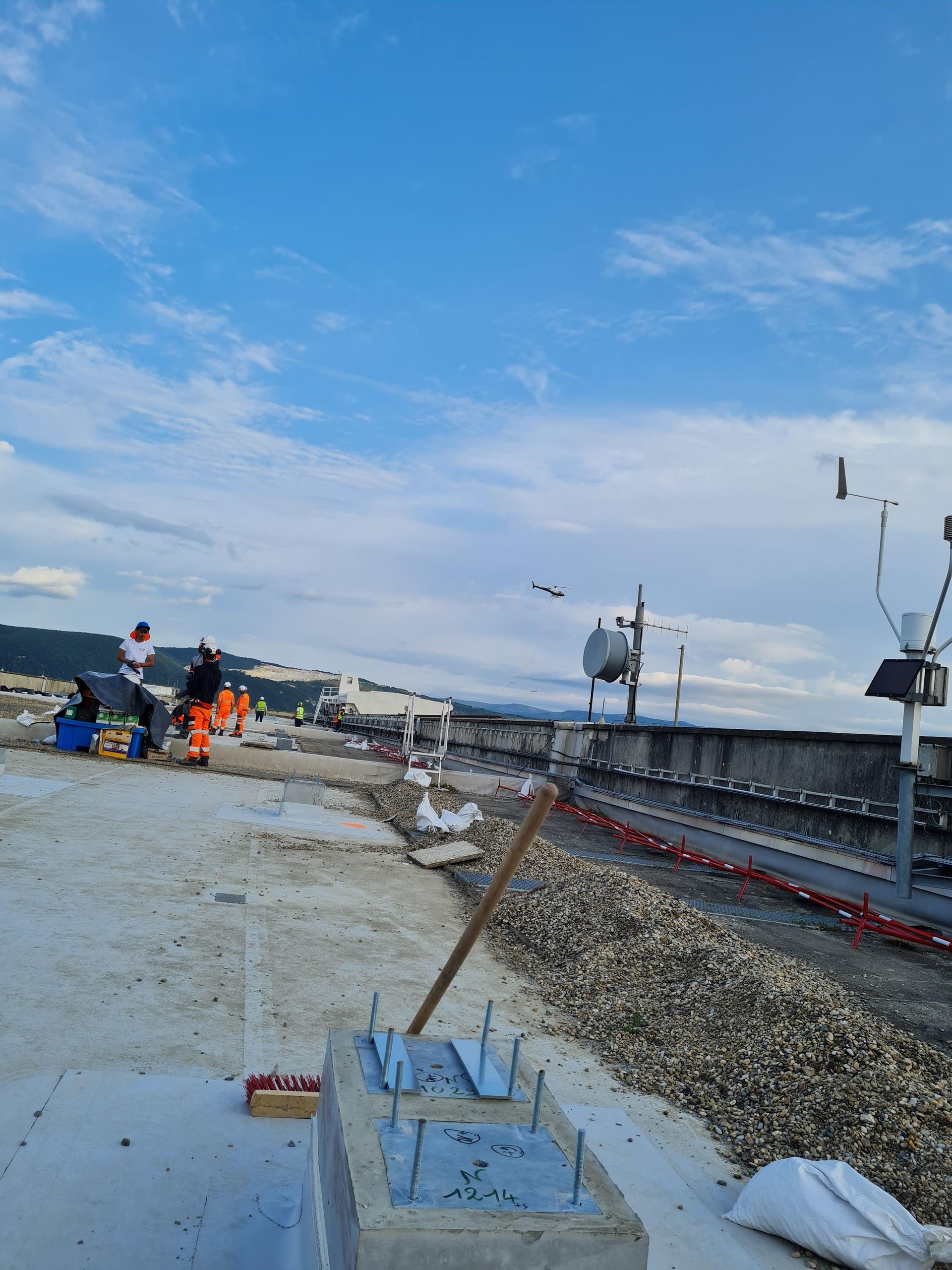 Des ouvriers sur un toit, près d'équipements et d'un muret, sous un ciel bleu nuageux.