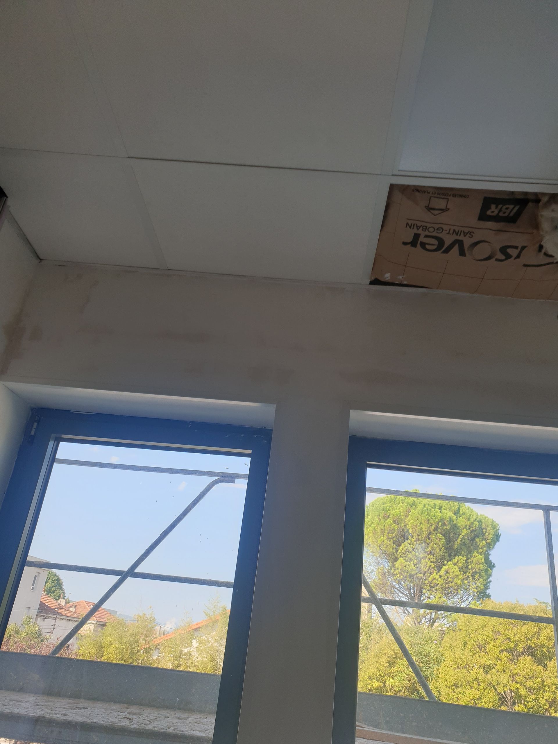 Vue intérieure d'une pièce avec fenêtres et plafond partiellement installé. On aperçoit des arbres verts et un ciel bleu à travers les fenêtres.