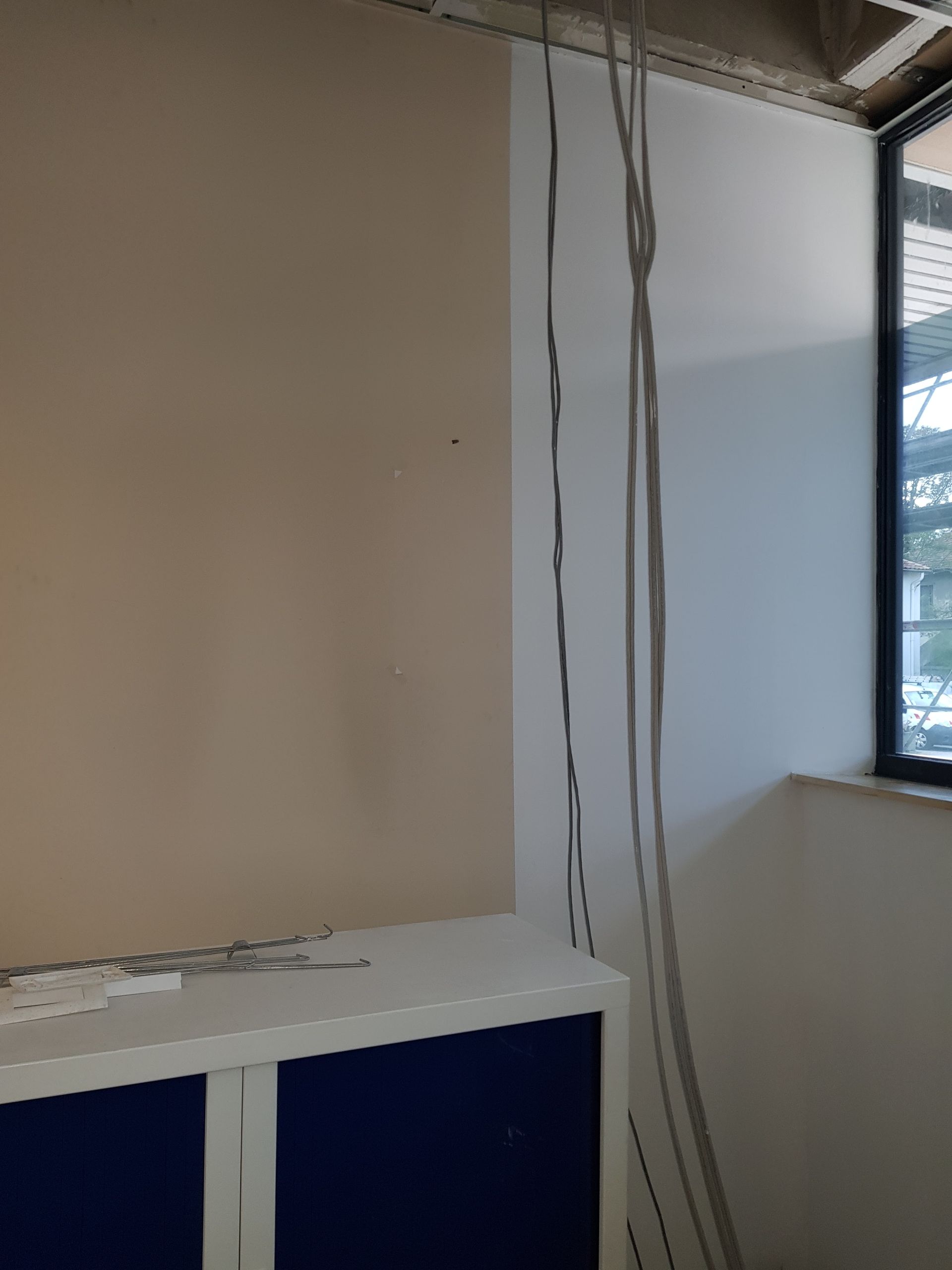 Des câbles courent le long d'un mur blanc, à côté d'un mur beige, au-dessus d'une armoire bleue et blanche, près d'une fenêtre.