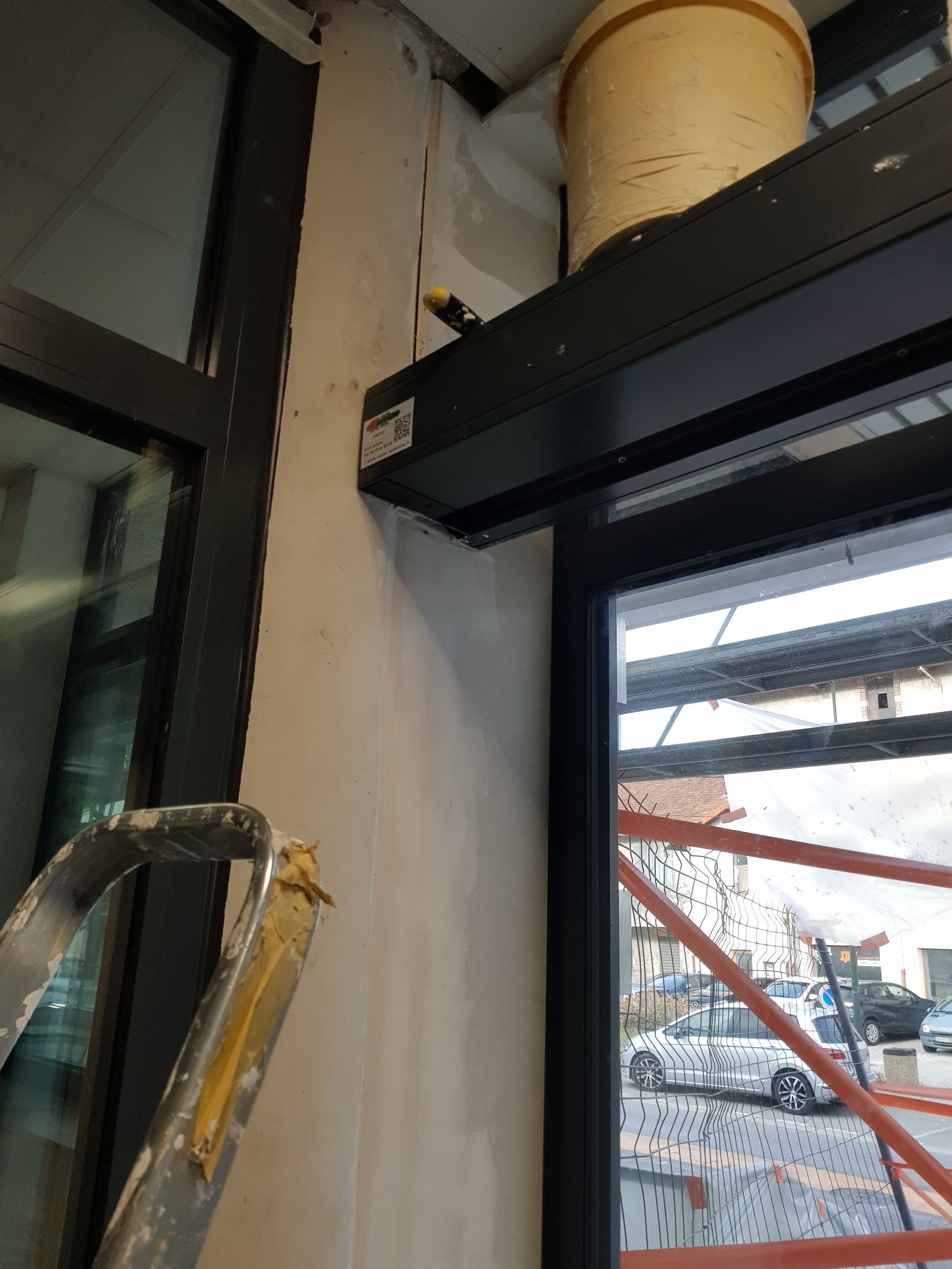 Échelle près d'une fenêtre et d'un mur ; mécanisme de volet au-dessus de la fenêtre ; scène extérieure à travers la fenêtre.