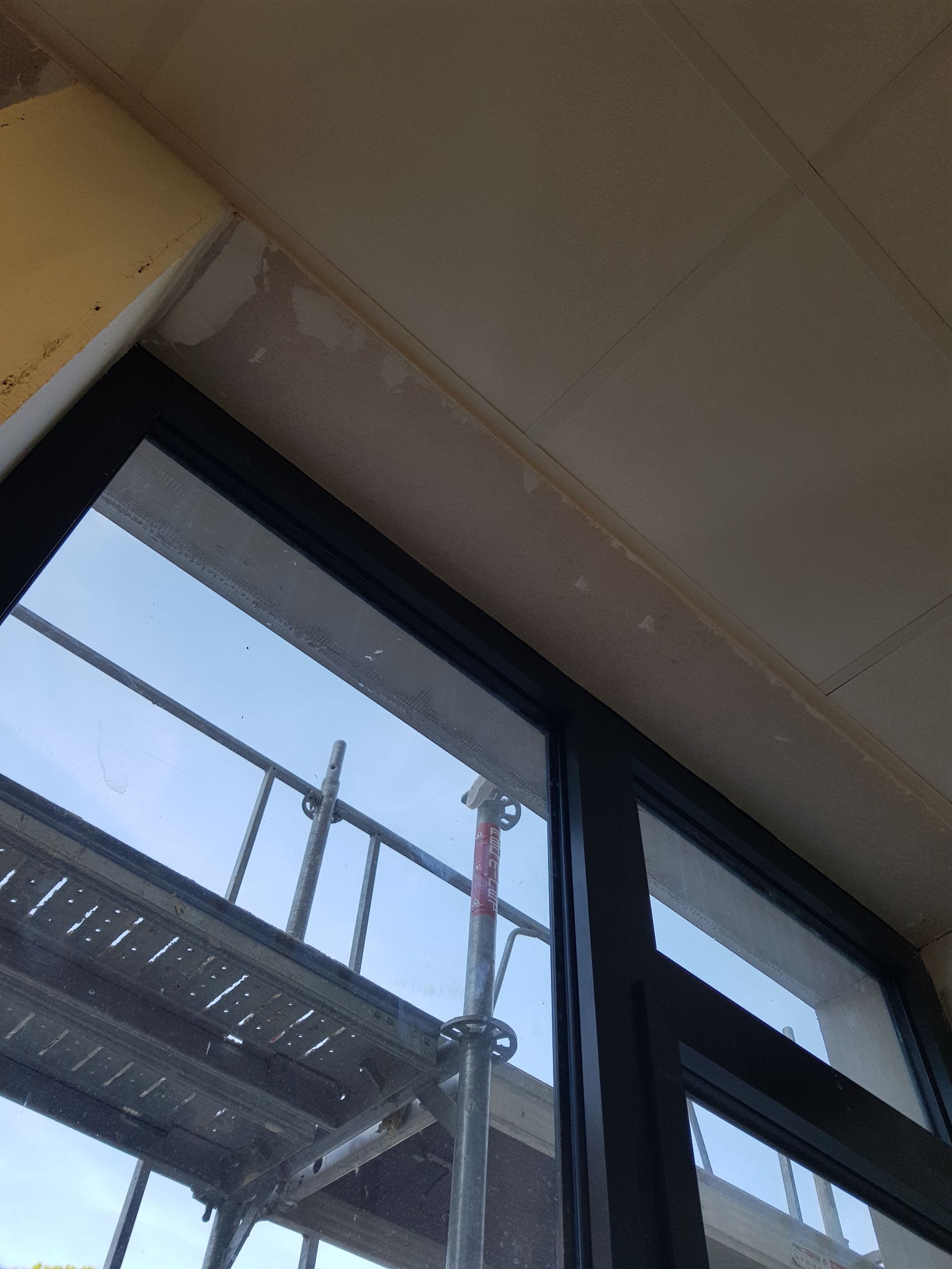 Fenêtre à cadre noir, plafond en plâtre inachevé, échafaudages se reflétant dans la vitre.