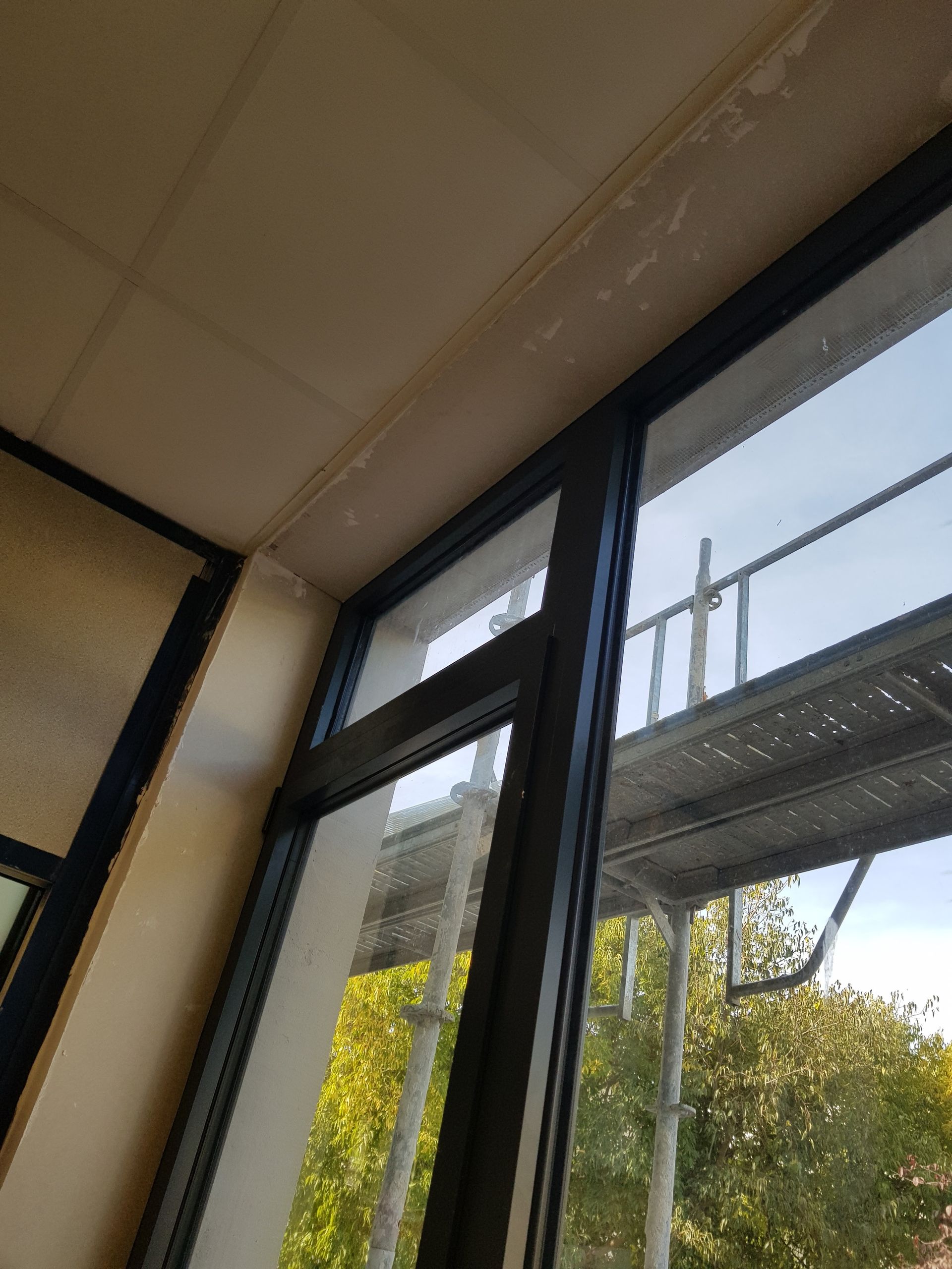 Fenêtre à cadre noir vue de l'intérieur d'une pièce. Échafaudages visibles à l'extérieur. Plafond blanc cassé.