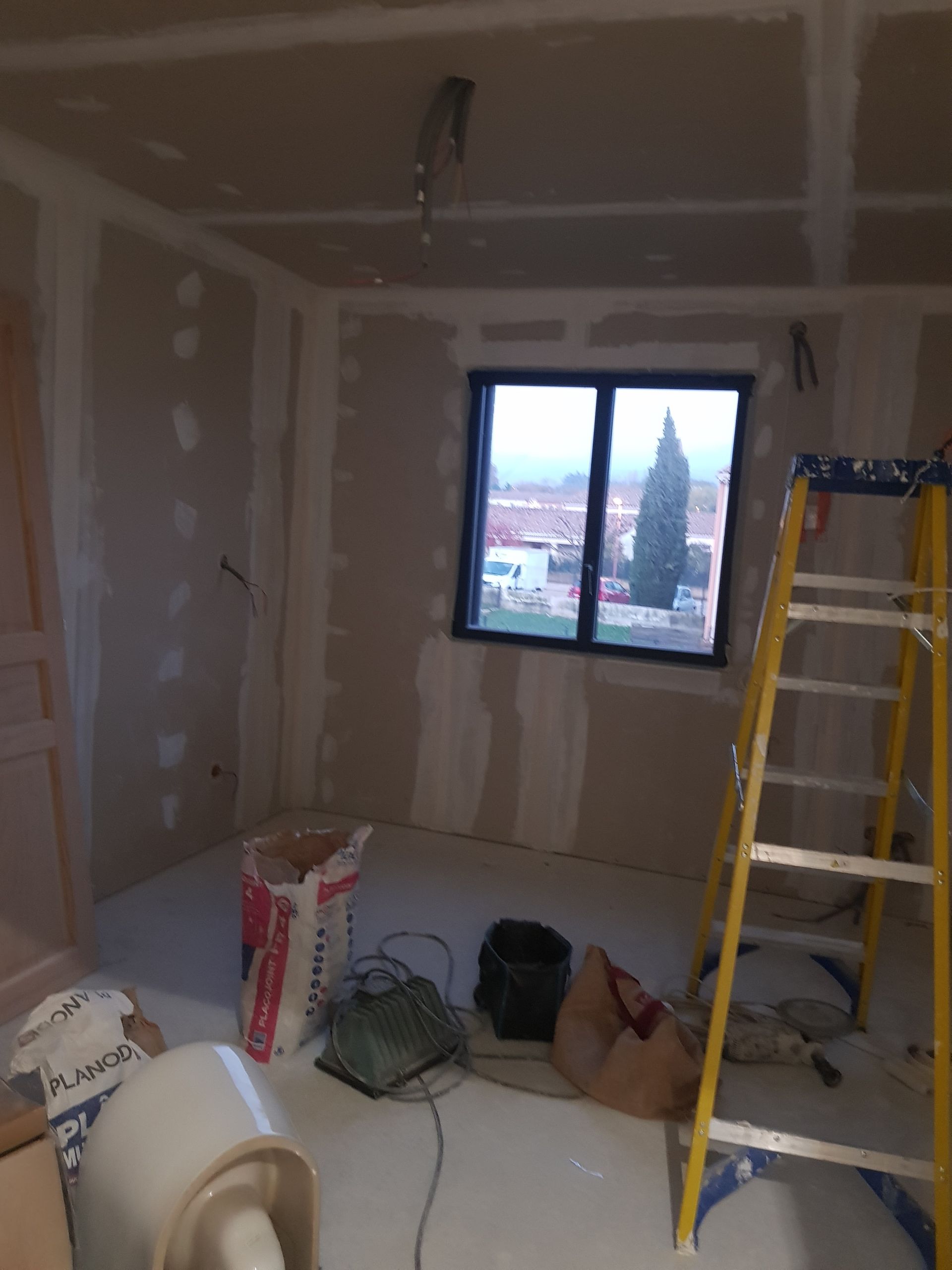 Pièce en travaux avec des cloisons sèches et une échelle près d'une fenêtre donnant sur la ville.
