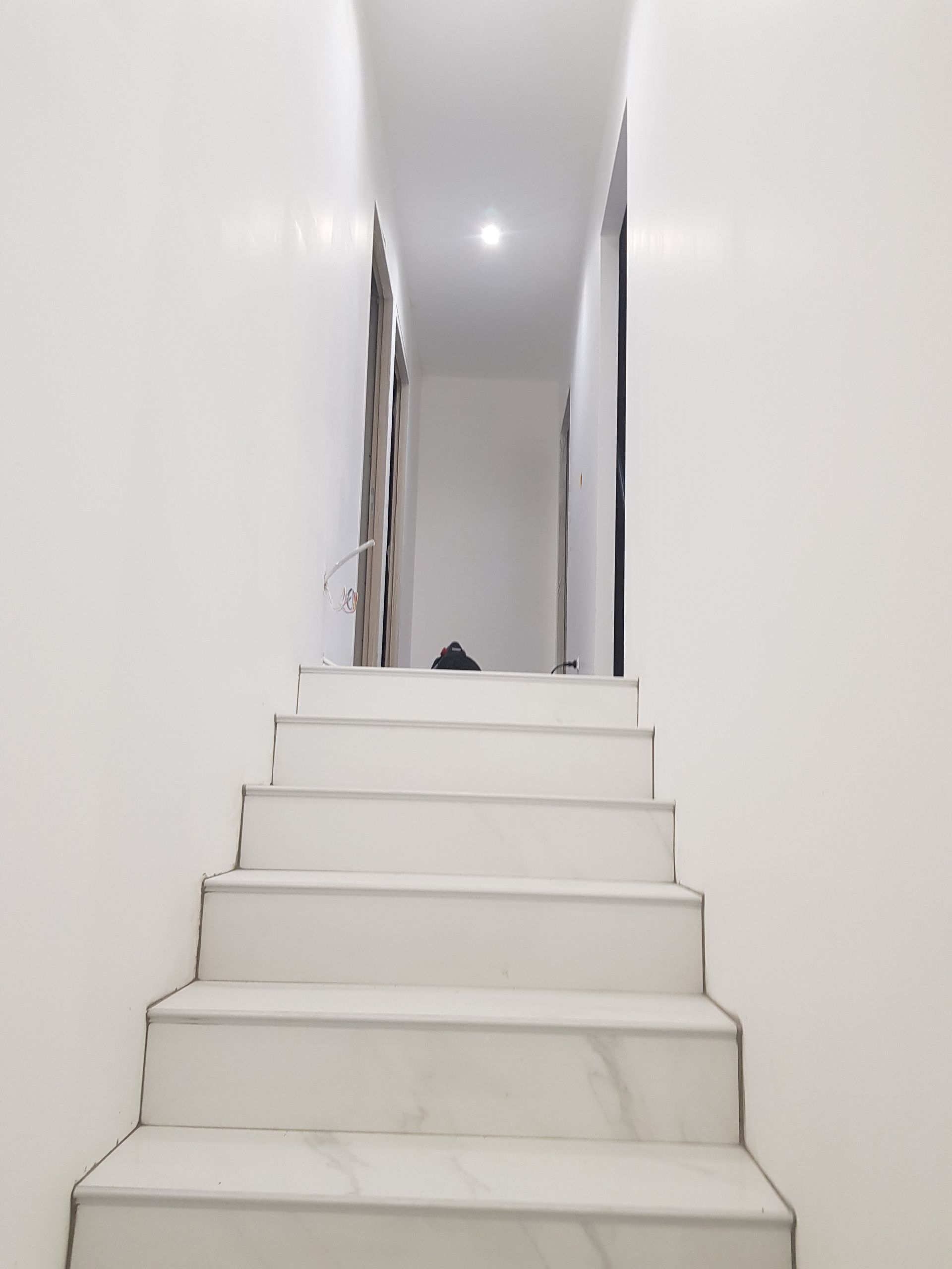Un escalier blanc mène à un couloir étroit éclairé au fond. Des murs blancs bordent l'escalier.