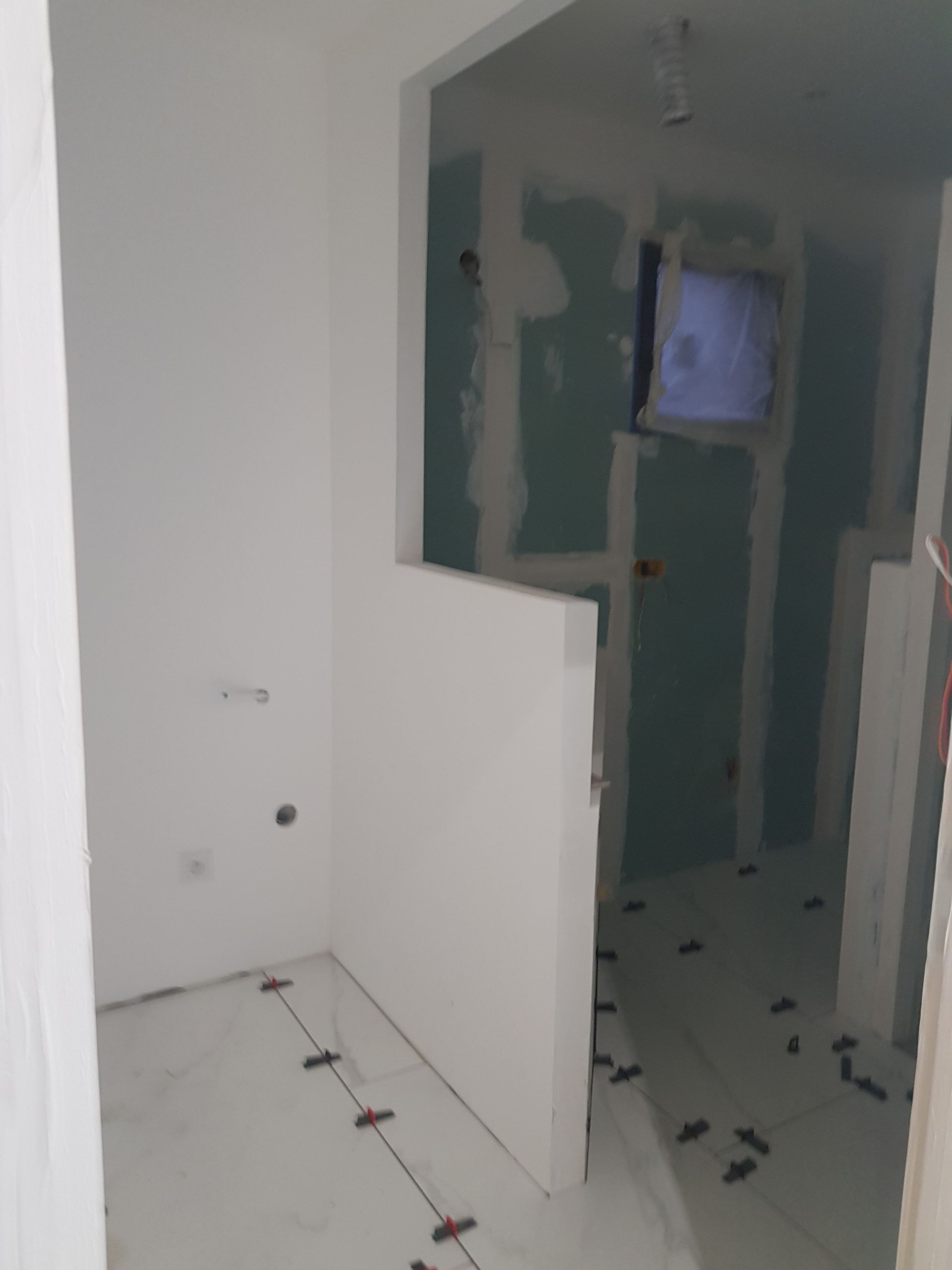 Intérieur en cours de construction : pièce aux murs blancs, cloisons sèches non finies et sol carrelé ; quelques outils sont présents.