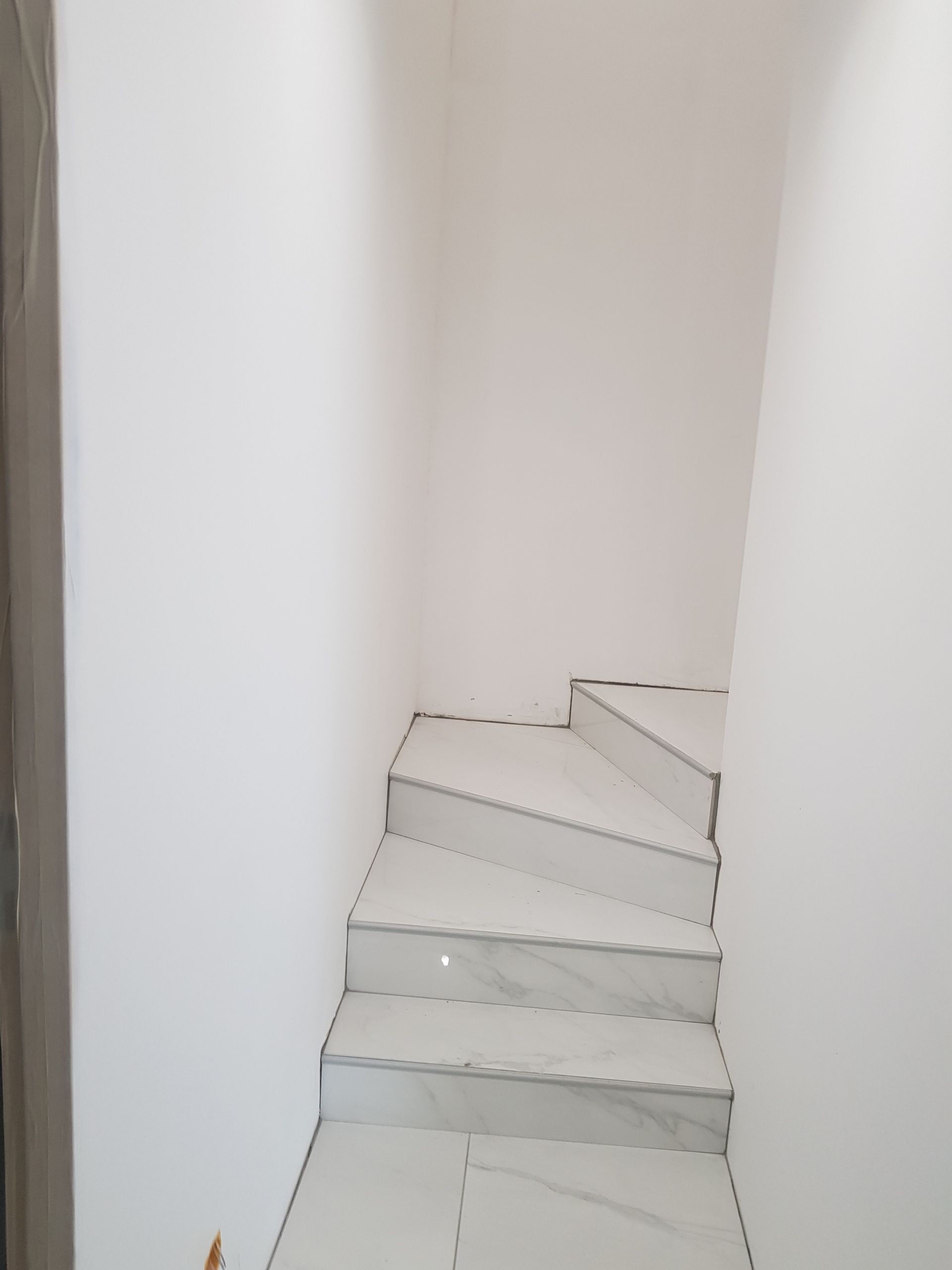 Un escalier carrelé de blanc mène à l'étage, entre des murs blancs.