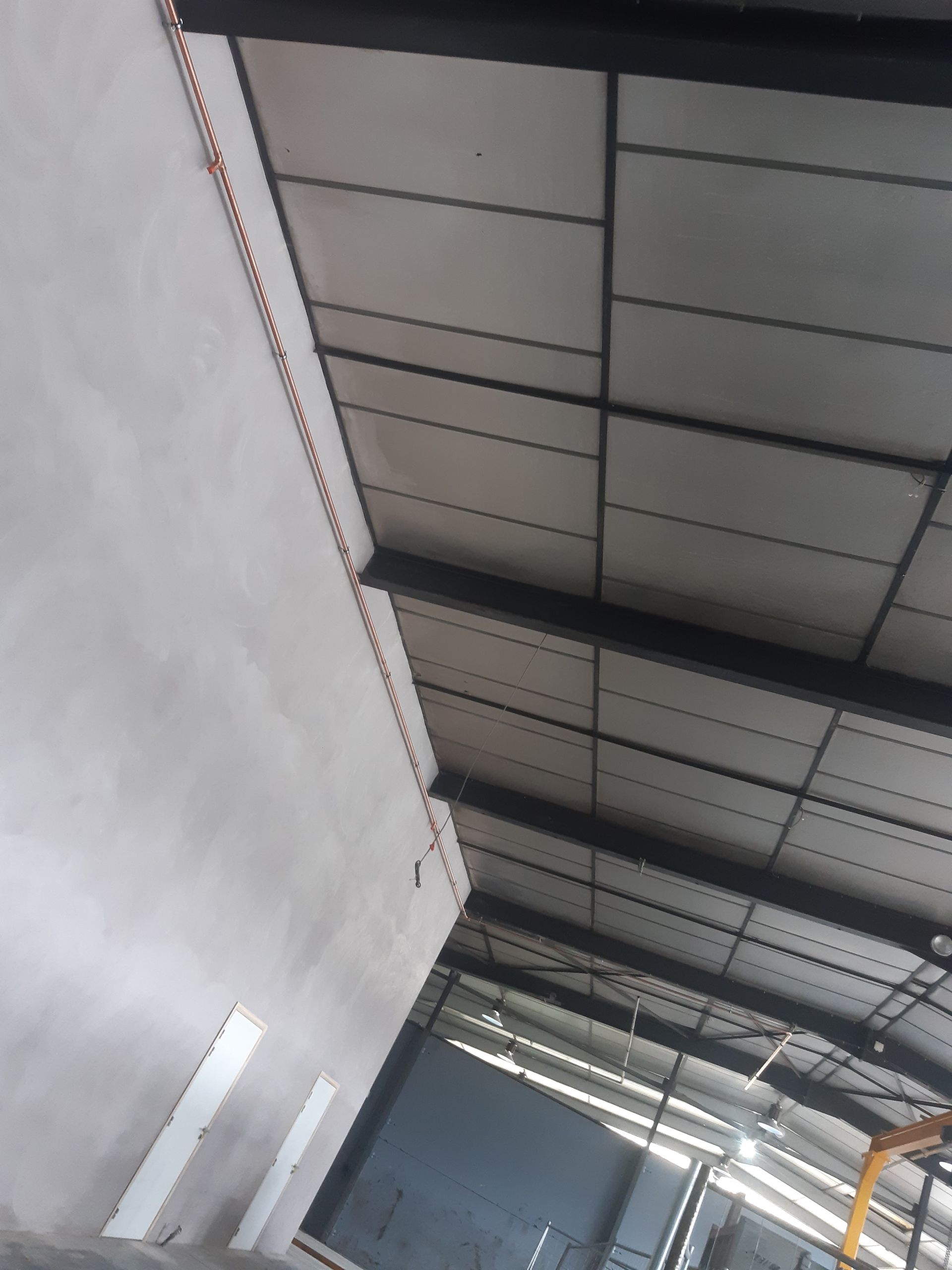 Intérieur d'un bâtiment industriel avec murs blancs, poutres en acier noir et panneaux de plafond gris.