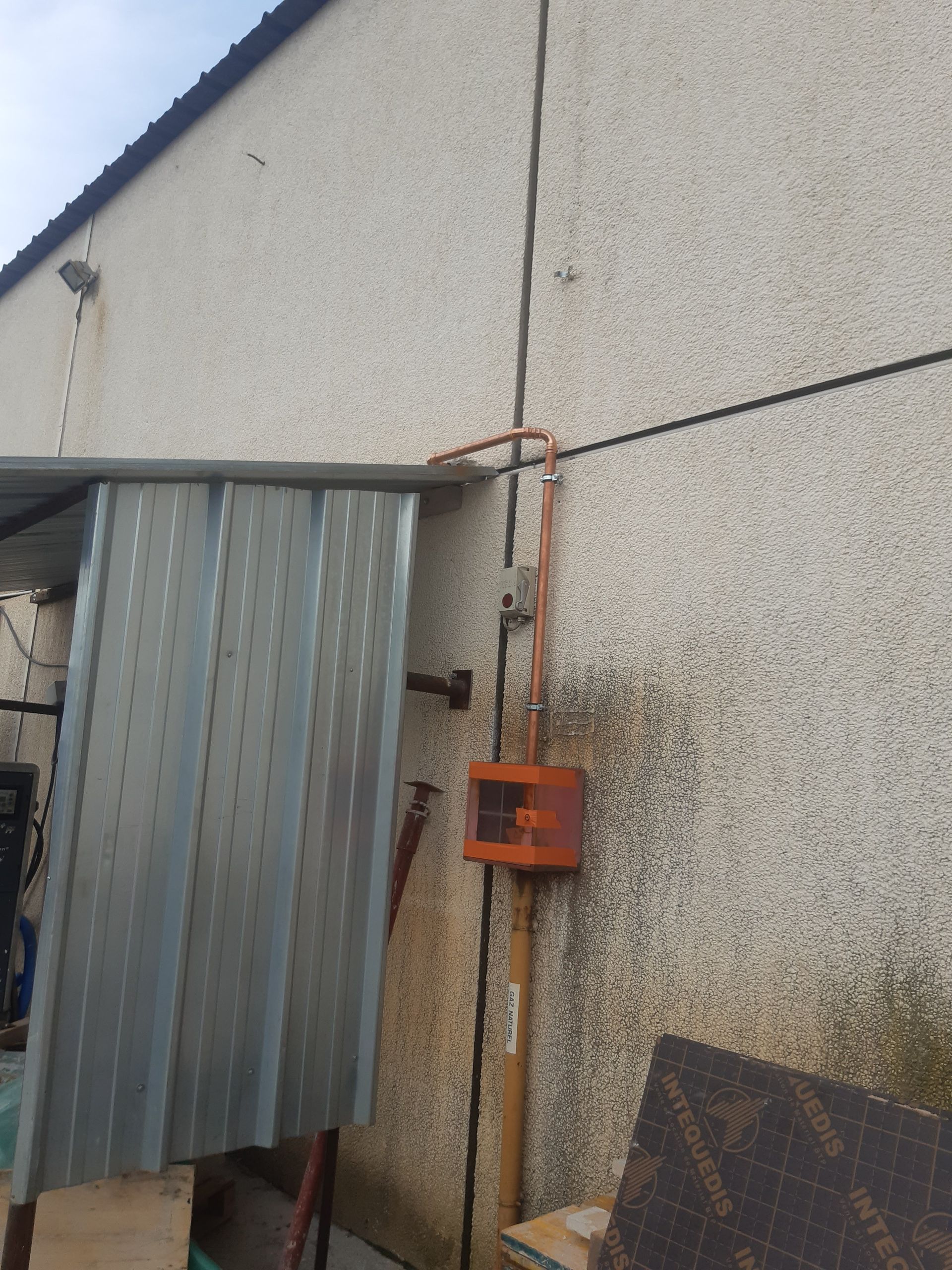 Vue extérieure d'un bâtiment avec un auvent en tôle ondulée. Un boîtier électrique et une gaine sont fixés à un mur texturé.