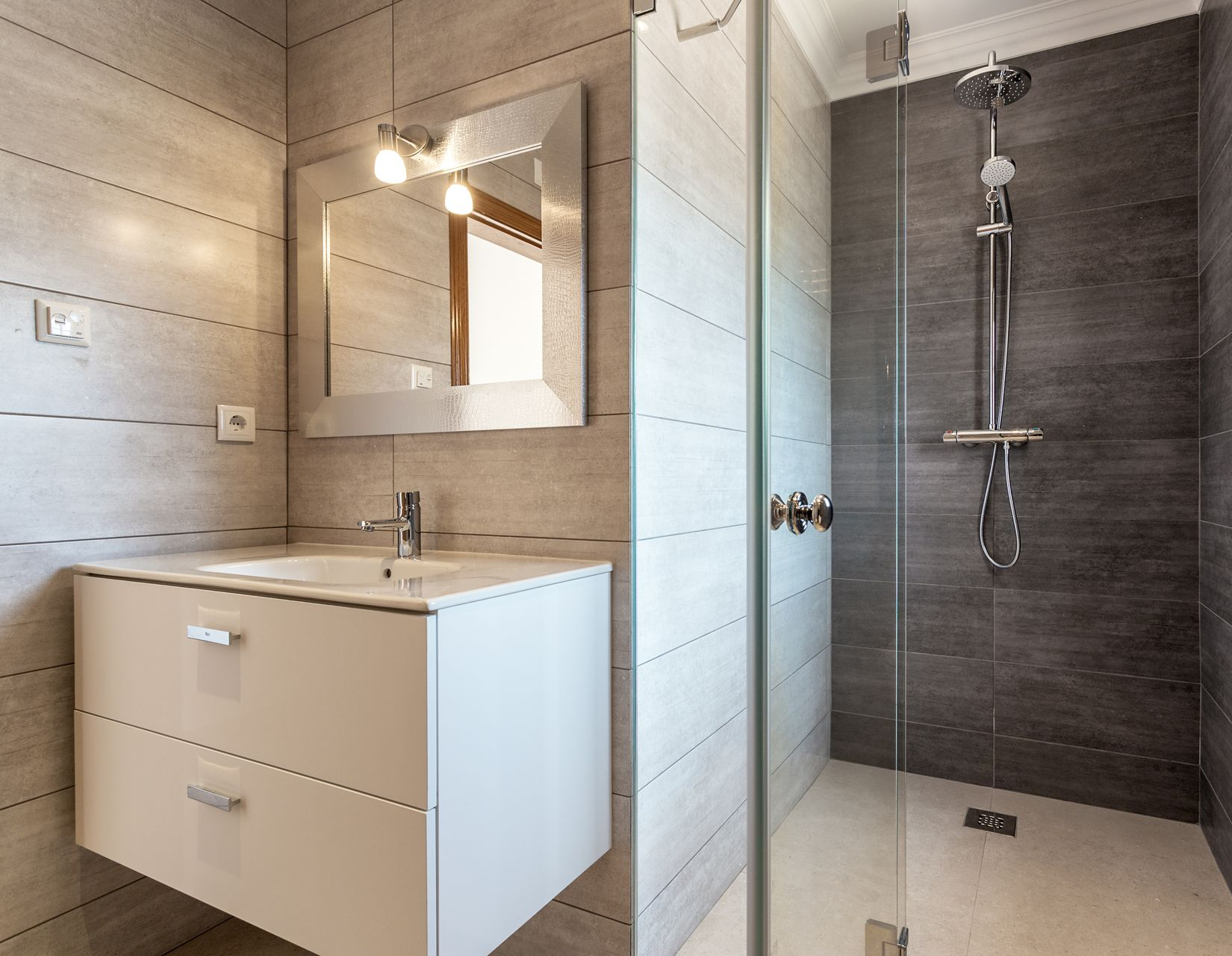 Salle de bains moderne avec murs carrelés gris, meuble-lavabo blanc, miroir et cabine de douche en verre.