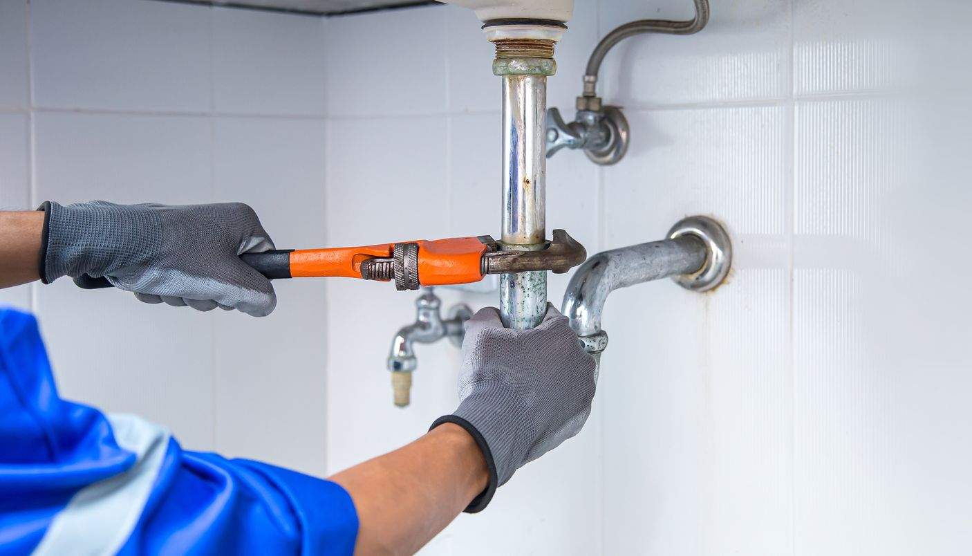 Un plombier portant des gants gris utilise une clé pour travailler sur des tuyaux sous un évier blanc.