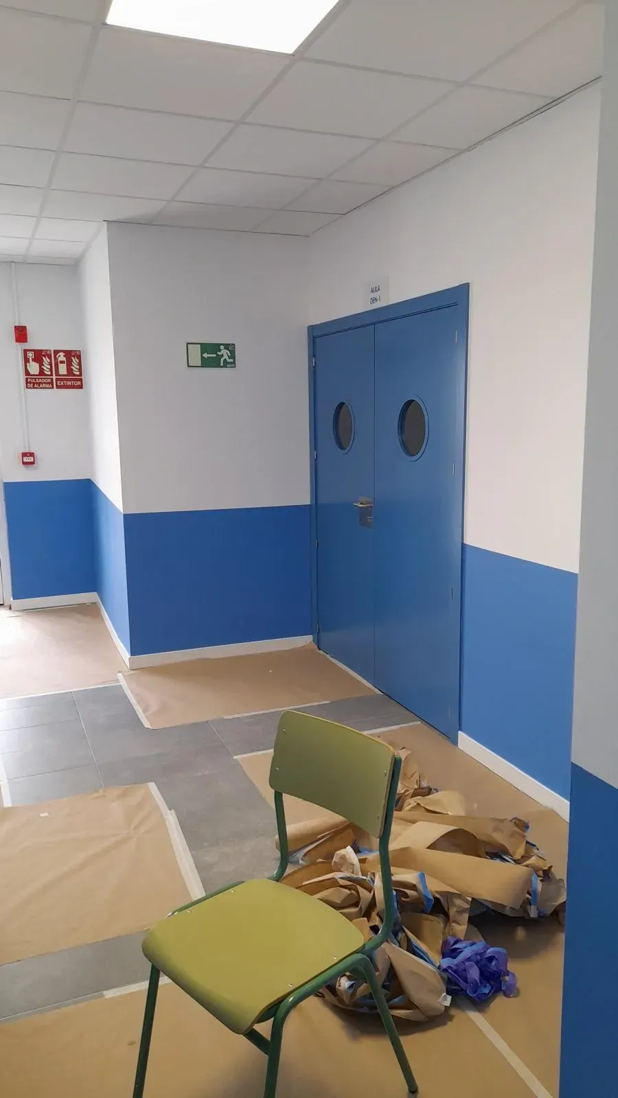 Pasillo con paredes azules y blancas, puertas dobles azules y sillas volcadas con escombros en el suelo.