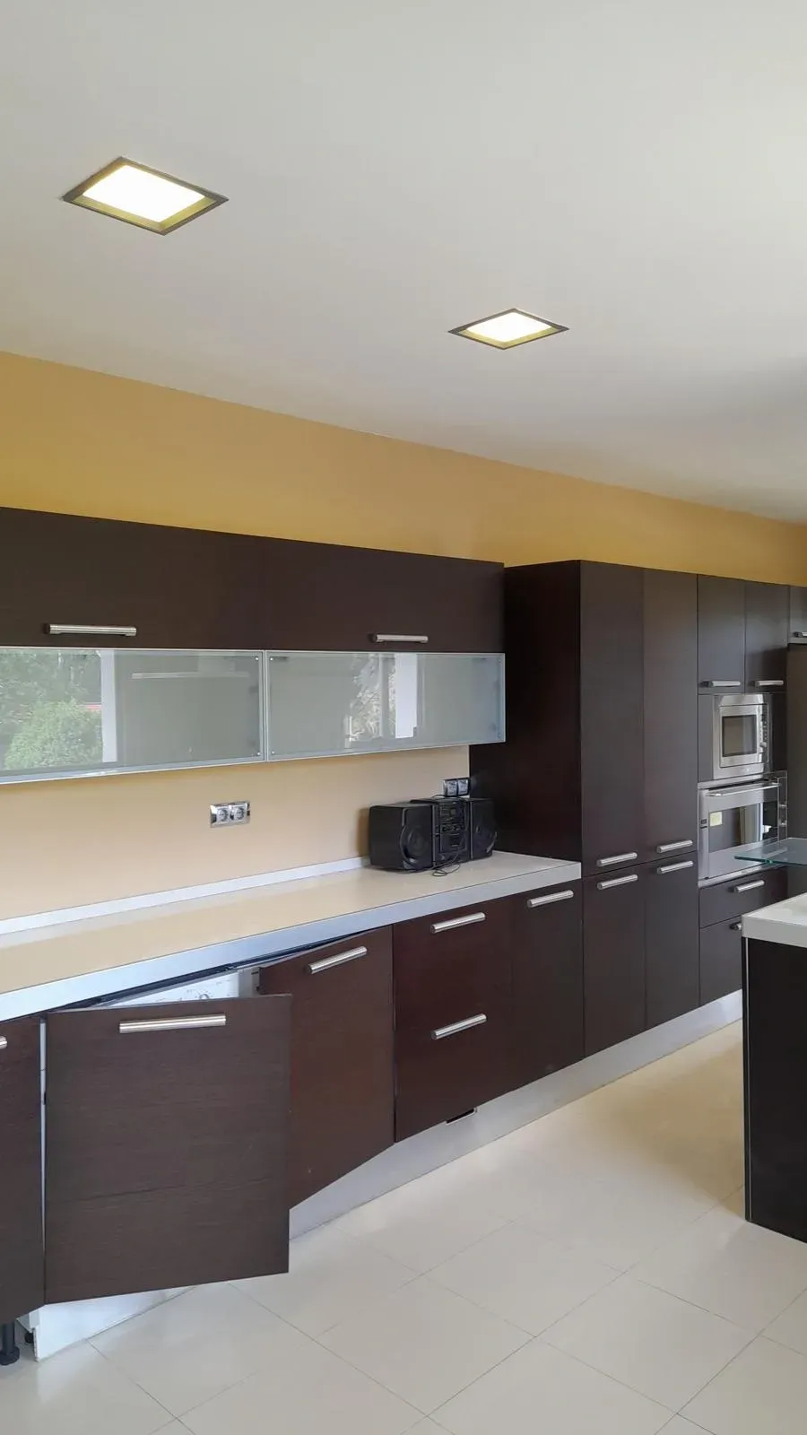 Cocina moderna con gabinetes oscuros, encimeras claras y electrodomésticos integrados bajo luces empotradas en el techo.