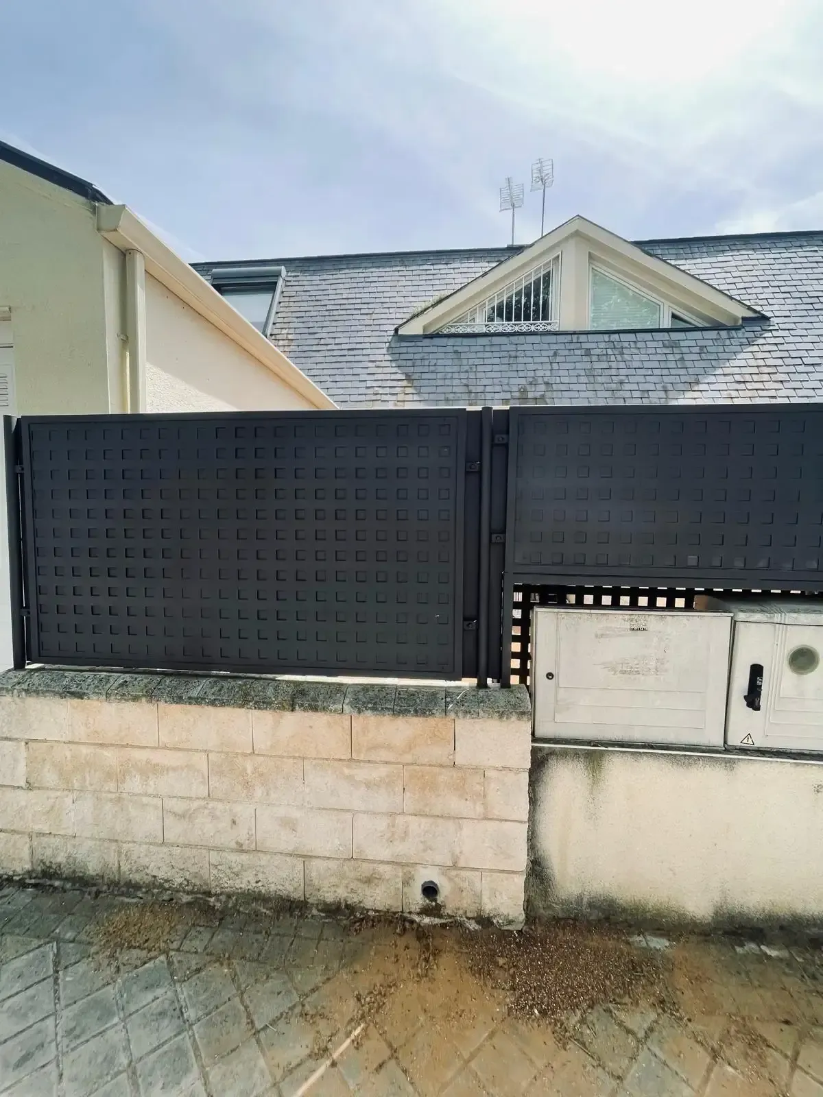 Una verja de metal negro y un muro bajo de piedra se encuentran frente a una casa con techo gris y buzón de correo.