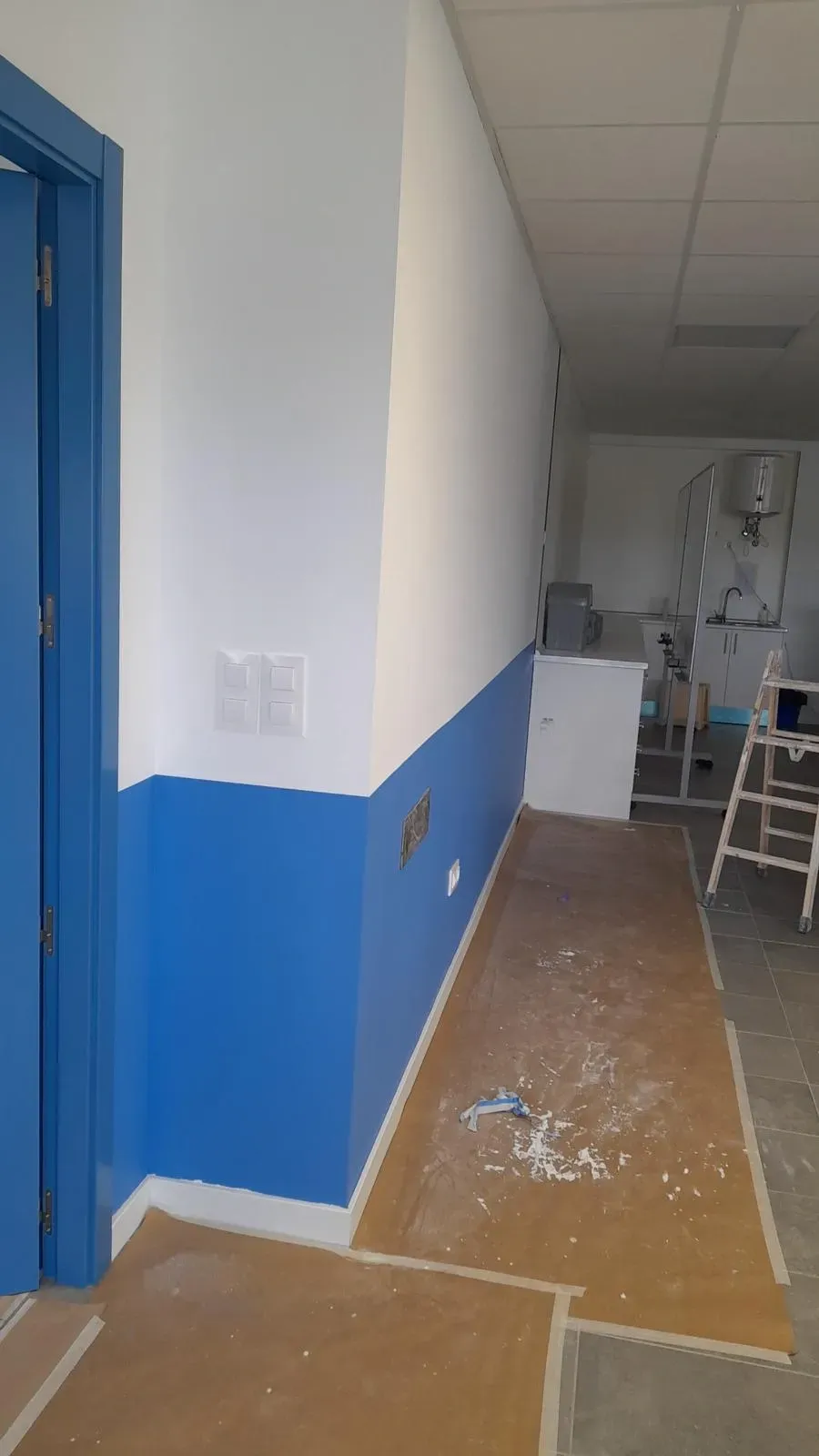 Pasillo estrecho con paredes azules y blancas, suelo desnudo y escombros de construcción en un espacio parcialmente renovado.