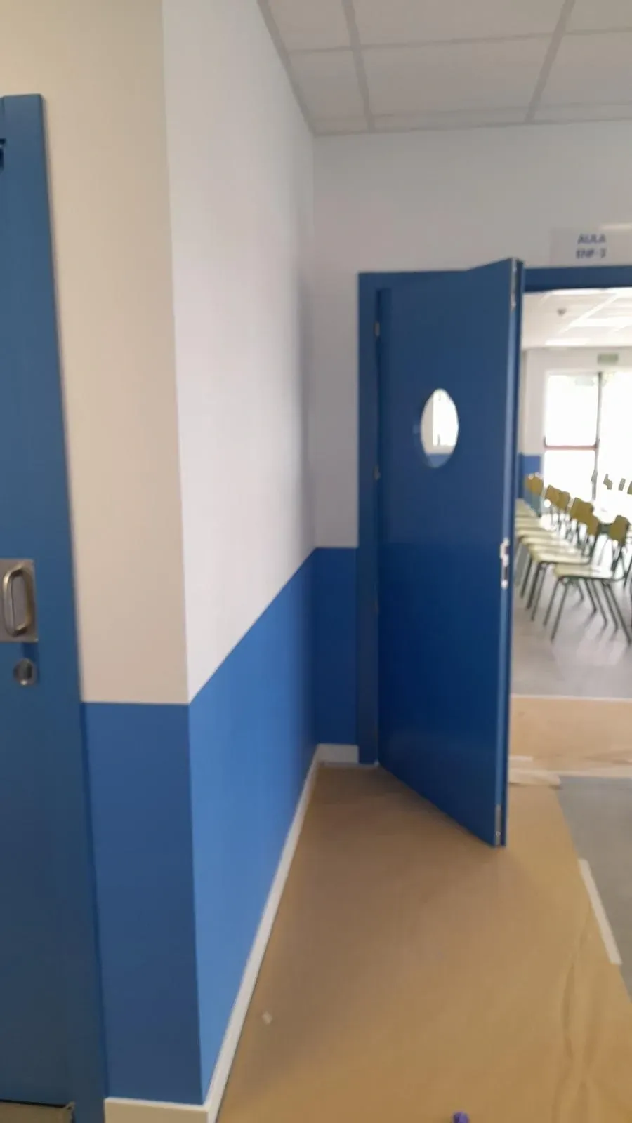 Pasillo azul y blanco con una puerta azul abierta que da a un aula con filas de sillas.