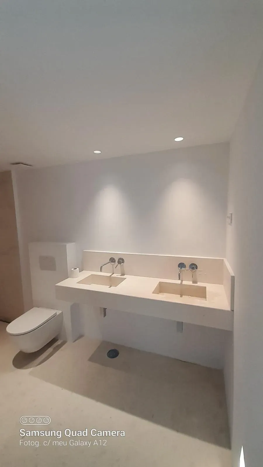 Baño minimalista blanco con inodoro suspendido y tocador con doble lavabo bajo luces empotradas.