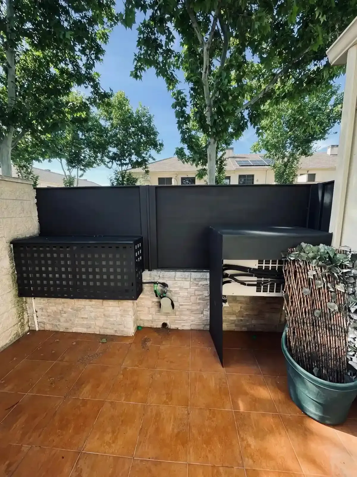 Patio exterior con baldosas de terracota, valla negra para mayor privacidad y una planta en maceta junto a una mesa negra.
