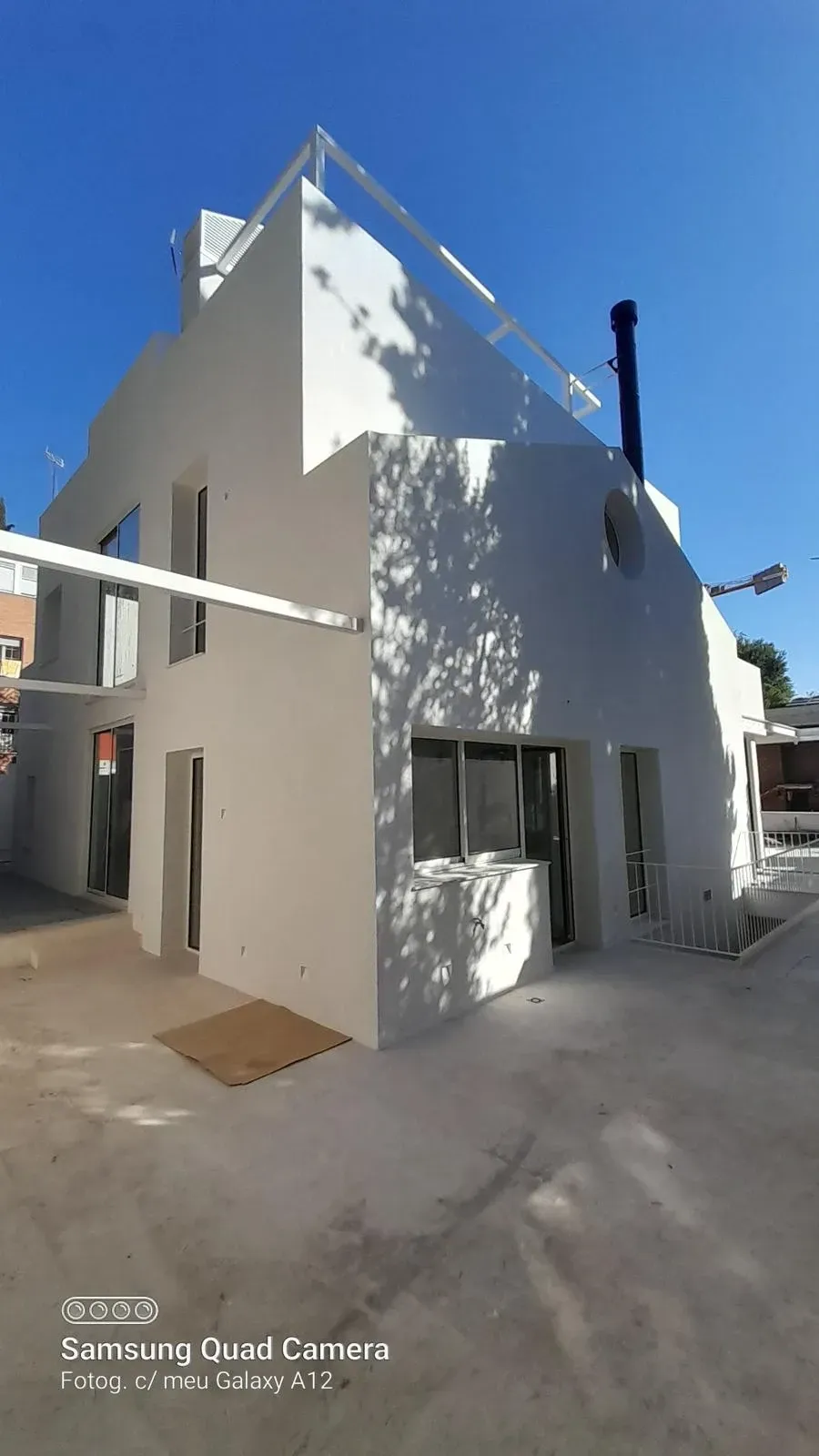 Edificio blanco en esquina con barandilla en la azotea, ventanas pequeñas y un callejón sombrío bajo un cielo azul brillante.