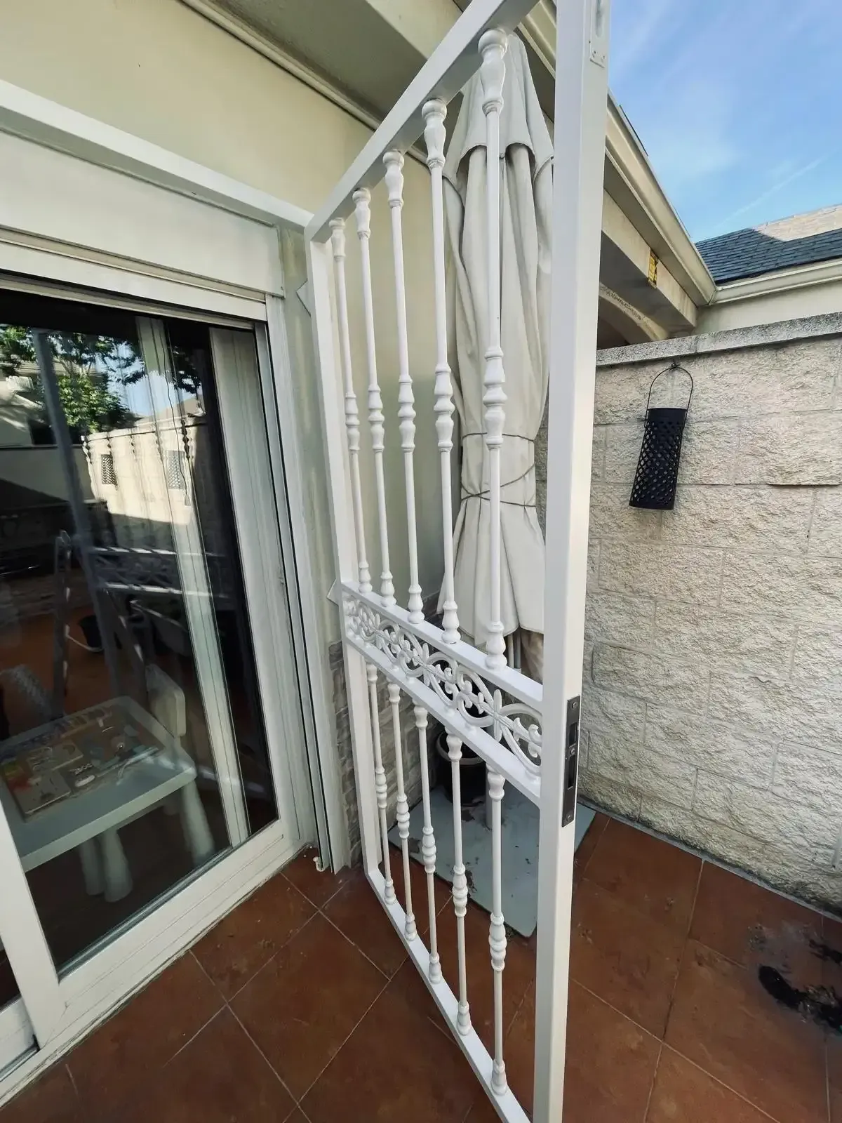 Una verja de seguridad de metal blanco se encuentra junto a una puerta corredera de cristal en un pequeño patio embaldosado.