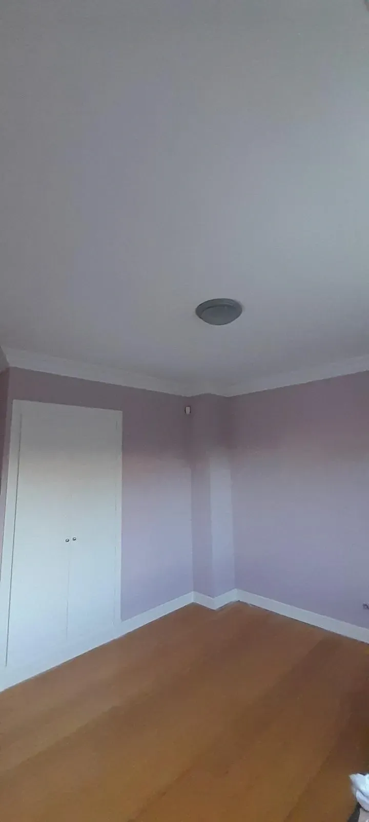 Rincón vacío de un dormitorio con paredes de color lila claro, puertas de armario blancas y suelo de madera.