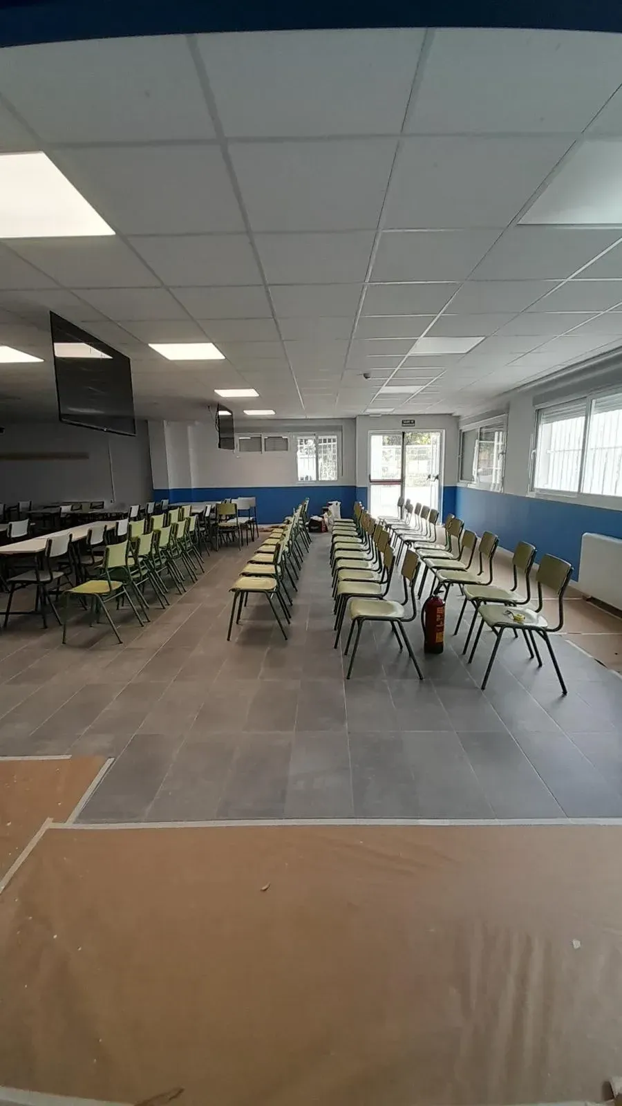 Cafetería vacía con filas de sillas y mesas verdes bajo luces brillantes en el techo.