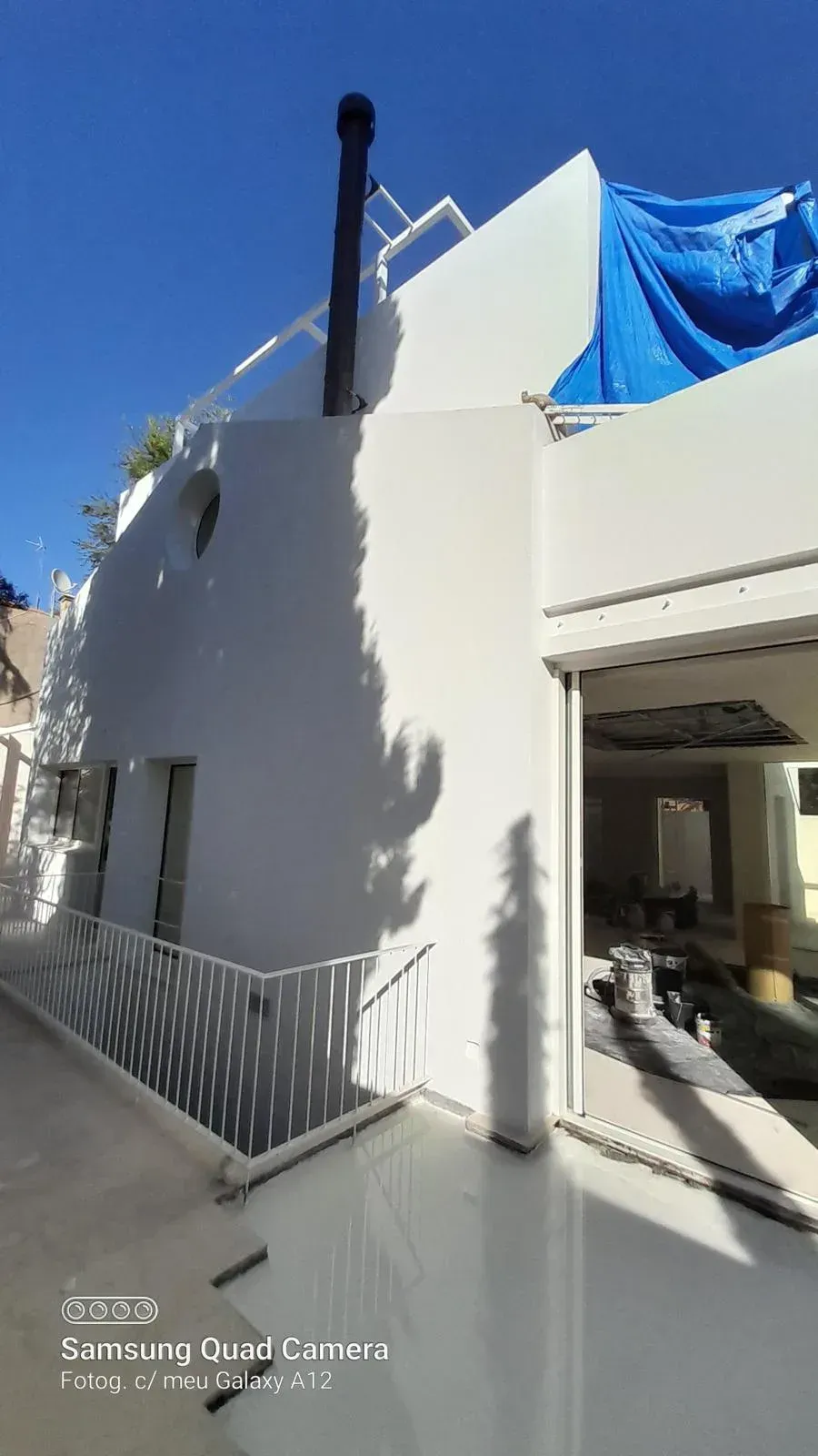 Edificio blanco en construcción con lona azul, barandilla metálica y luz solar brillante que proyecta sombras.