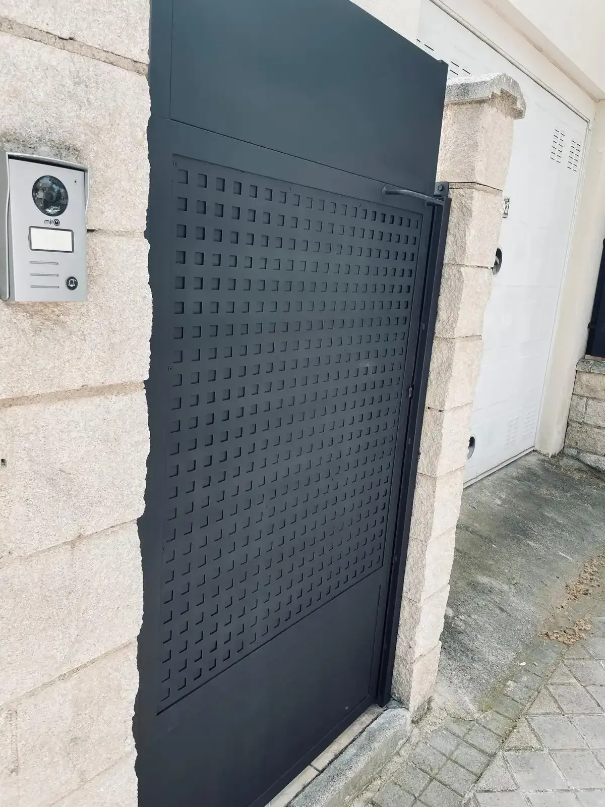 Una puerta peatonal de metal negro, con un pequeño panel de intercomunicación al lado, se encuentra sobre un muro de piedra.