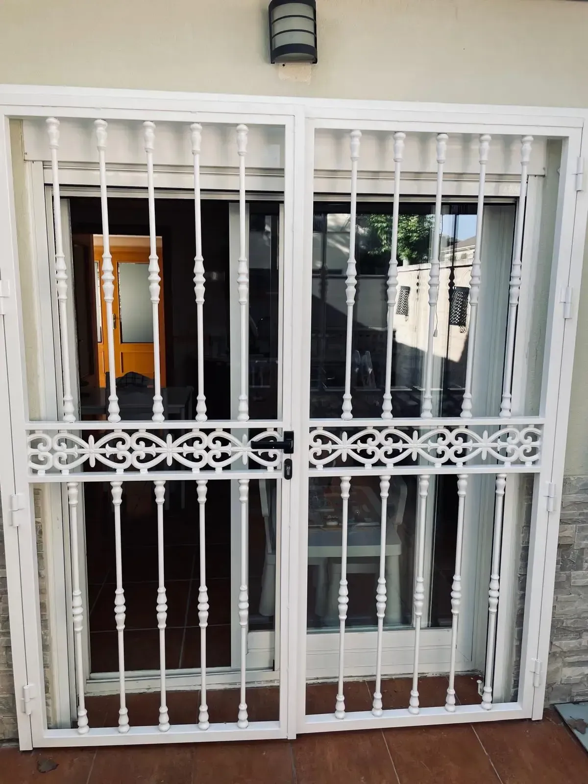 Puerta de seguridad de metal blanco con barrotes verticales y celosía decorativa delante de una puerta y una ventana.