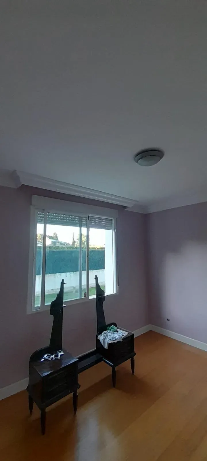 Habitación vacía con dos sillas negras junto a una ventana, paredes de color lila claro y suelo de madera.