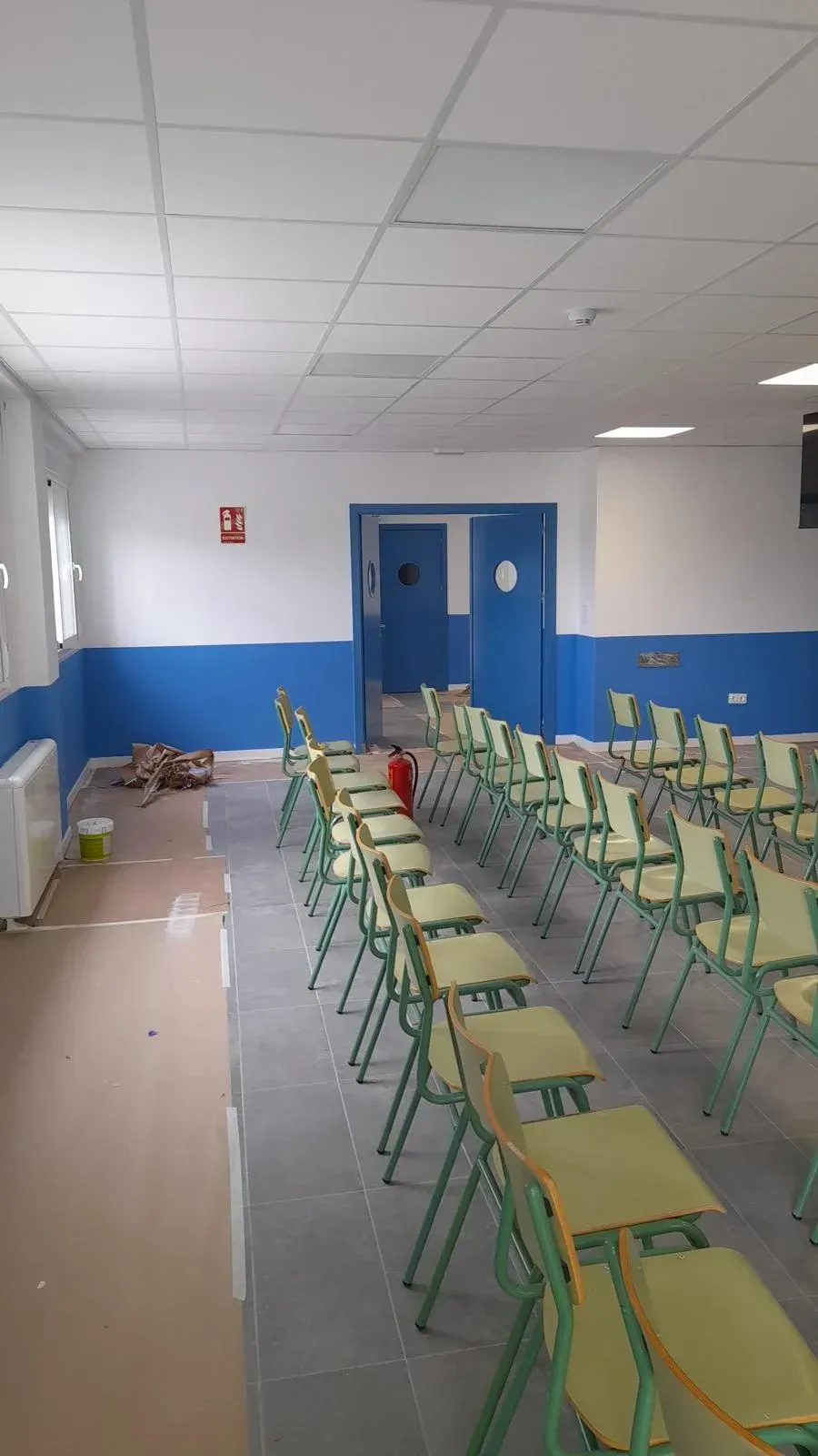 Aula vacía con filas de sillas y pupitres verdes, paredes azules y blancas, y puertas dobles al fondo.