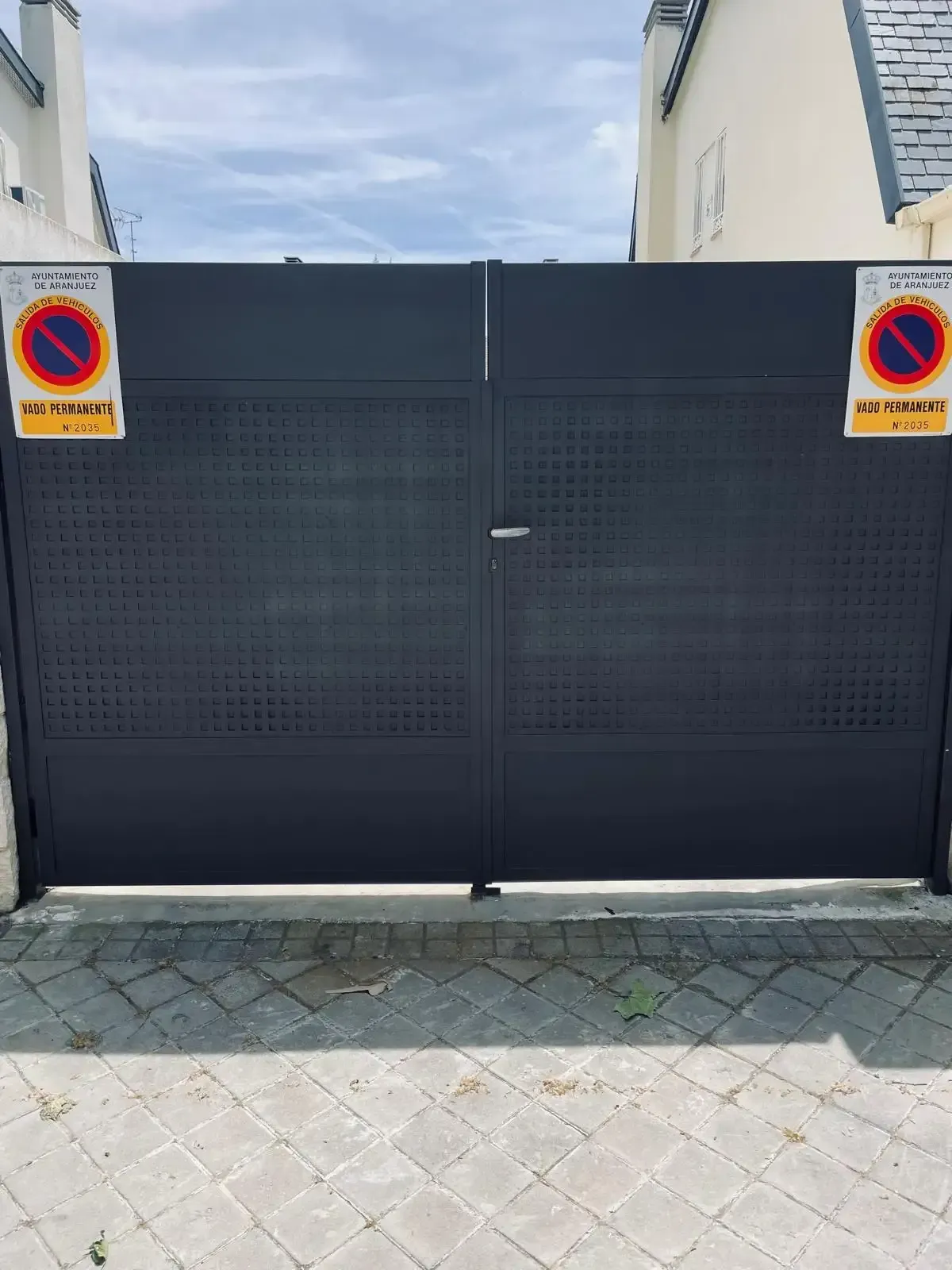 Una puerta metálica negra con señales de prohibido aparcar a ambos lados, vista desde un camino de entrada pavimentado.