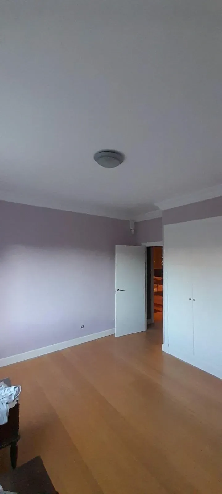 Habitación vacía con suelo de madera clara, paredes pálidas, puertas blancas y lámpara de techo.