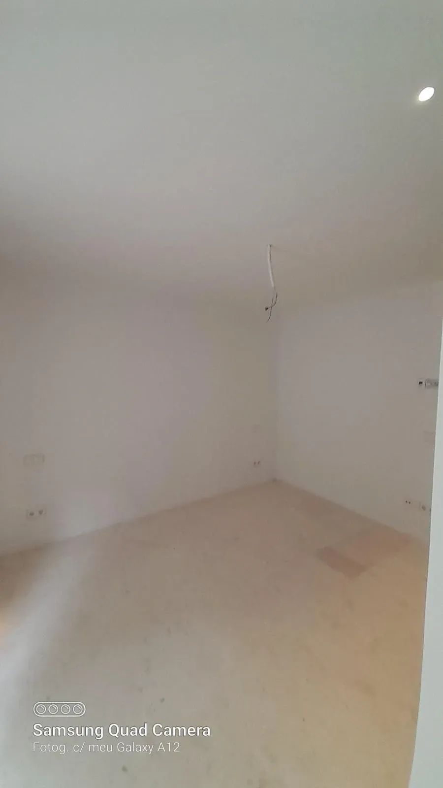 Habitación blanca vacía con suelo beige y un único cable de lámpara colgante del techo.